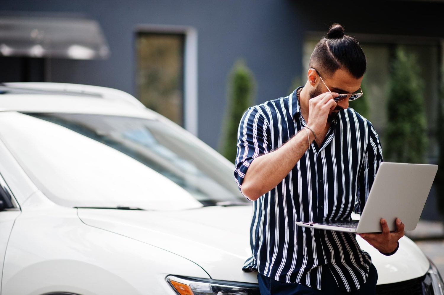 Man in a striped shirt leaning on a white SUV while researching how to pawn a car with outstanding finance on his laptop